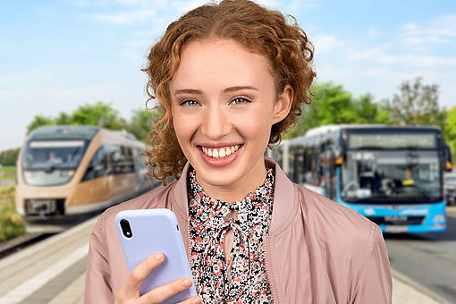 Eine junge Frau mit Smartphone steht an einem Bahnhof vor einem Zug und einem Bus.